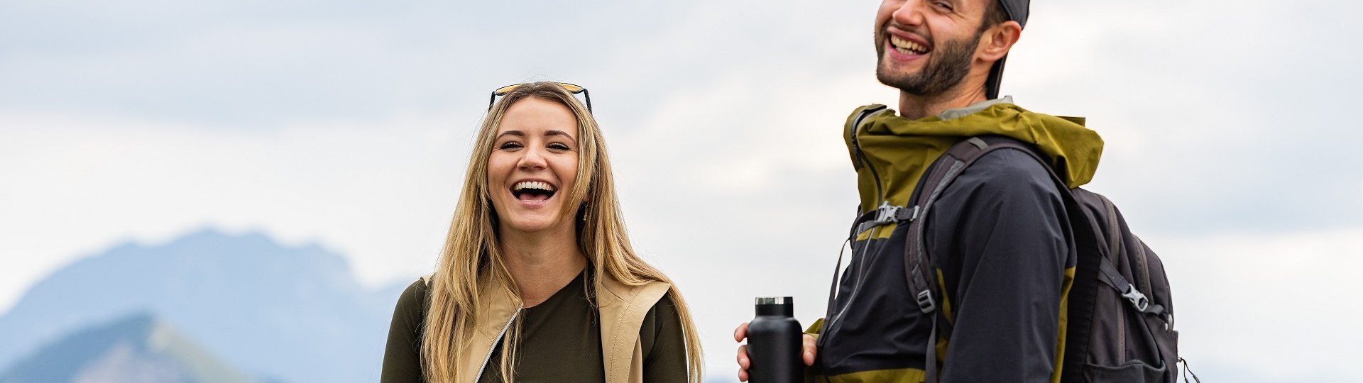 Lachend stehen eine junge Frau und ein junger Mann in Wanderkleidung auf einem Berg und machen eine kurze Pause zum Trinken., © Tölzer Land Tourismus|Farmhouse7