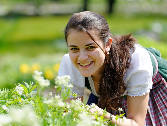 Eine im Dirndl gekleidete junge Frau beugt sich zu wei&szlig;en Bl&uuml;ten., &copy; G&auml;steinformation Bad Heilbrunn