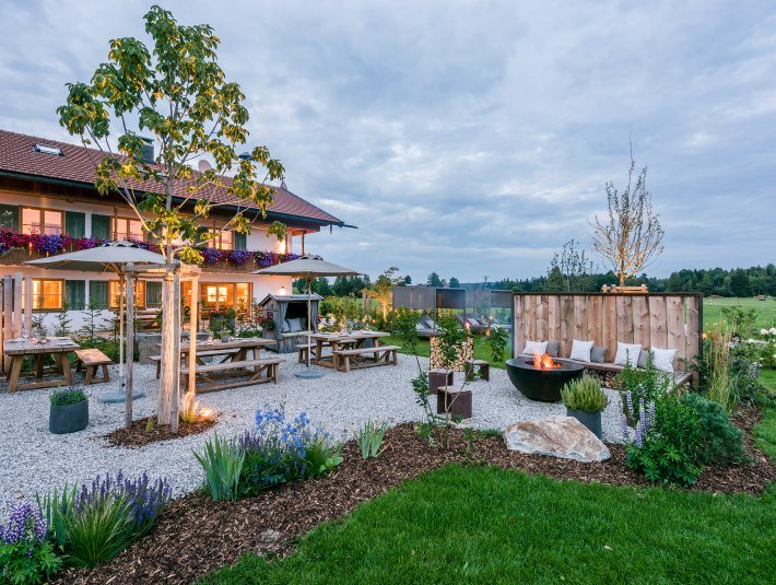 Das Bild zeigt den Außenbereich mit des Urlaub auf dem Bauernhof - Anbieters in abendlicher Stimmung mit Sonnenschirmen und Gartenlounge-Stühlen und eine große, runde Metall-Feuerstelle, © Hussnhof Foto: Günter Standl