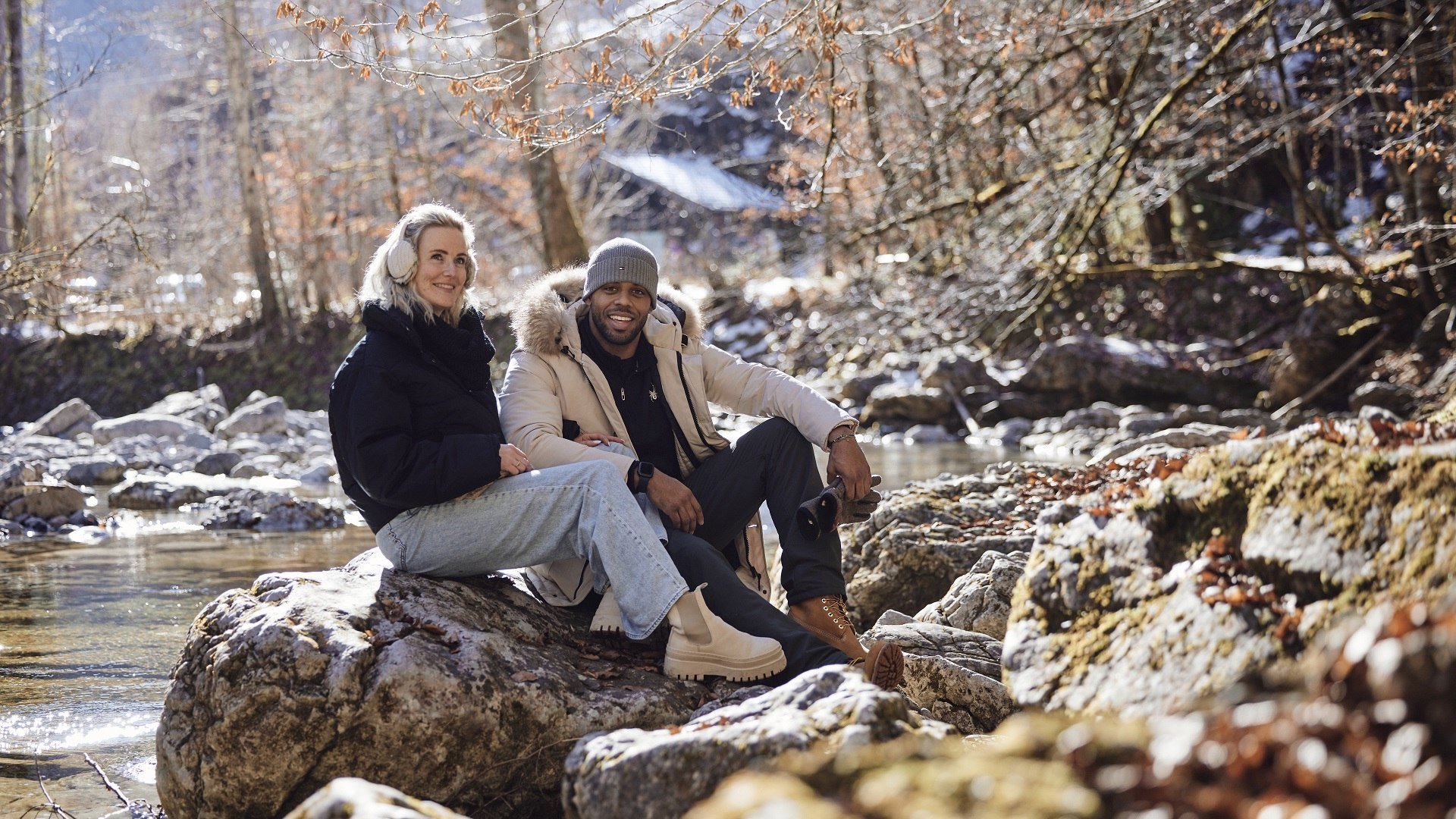 Zwei Personen sitzen entspannt auf einem großen Stein im Fluss, umgeben von Bäumen und Blättern im Winter und genießen sichtlich die herbstlich-winterliche Atmosphäre., © oberbayern.de|Tobias Köhler