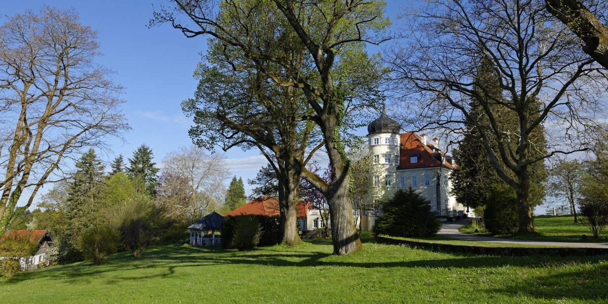 In der gr&uuml;nen Parkwiese mit gro&szlig;en B&auml;umen ist im Bild rechts die zart-gelbe Parkvilla mit wei&szlig;en Fenstern, rundem Turm und rotem Dach zu sehen. Dar&uuml;ber leuchtet der klare blaue Himmel., &copy; Lisa Bahnm&uuml;ller