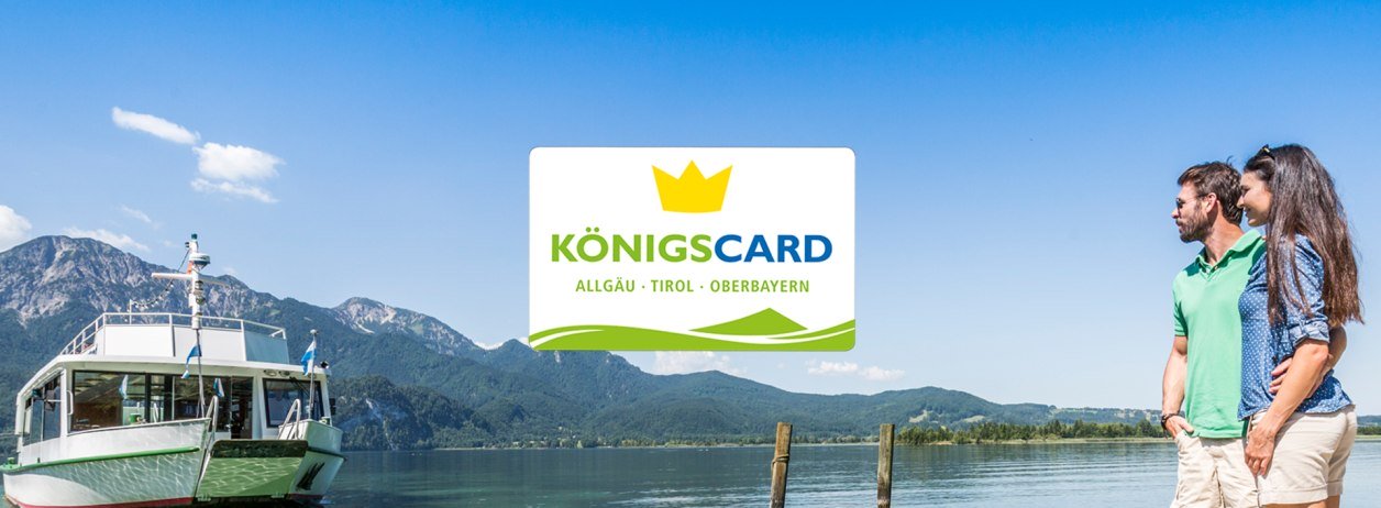 Die K&ouml;nigscard - bei ausgew&auml;hlten Gastgebern erh&auml;ltlich f&uuml;r ein MEHR an Urlaubsspa&szlig;. Das Bild zeigt ein auf das Kochelseeschiff wartende P&auml;rchen am Steg mit dem Logo der K&ouml;nigscard G&auml;stekarte, &copy; Foto: TI Kochel a. See|Thomas Kujat