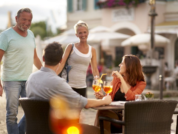 Bars &amp; Cafés verströmen südländisches Flair in der Isarstadt Bad Tölz.  Das Bild zeigt: Zwei Personen sitzen draußen an einem Tisch und stoßen mit Getränken an, zwei weitere Personen gehen im Hintergrund vorbei., © Tölzer Land Tourismus|Jan Greune