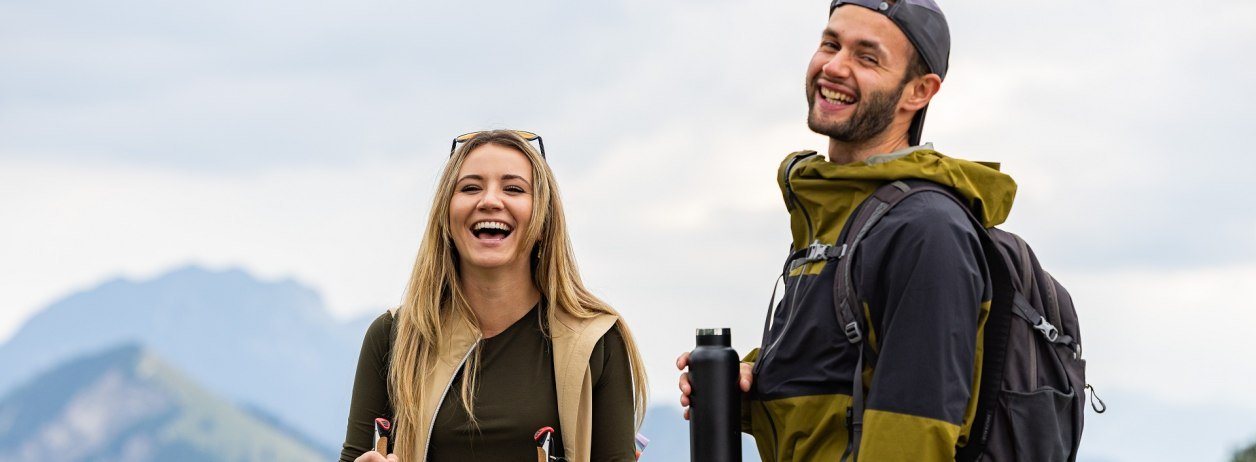 Lachend stehen eine junge Frau und ein junger Mann in Wanderkleidung auf einem Berg und machen eine kurze Pause zum Trinken., © Tölzer Land Tourismus|Farmhouse7