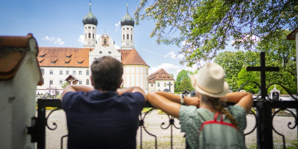 Auf dem Bild sind zwei Personen von hinten zu sehen, die an einem schmiedeeisernen Zaun lehnen und auf das Kloster Benediktbeuern mit seinen beiden markanten Zwiebelt&uuml;rmen blicken. Es ist ein sonniger Tag mit blauem Himmel und gr&uuml;nen B&auml;umen., &copy; T&ouml;lzer Land Tourismus, Deutsche Alpenstra&szlig;e|Dietmar Denger