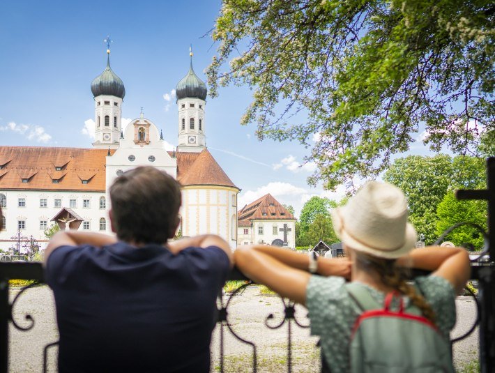 Auf dem Bild sind zwei Personen von hinten zu sehen, die an einem schmiedeeisernen Zaun lehnen und auf das Kloster Benediktbeuern mit seinen beiden markanten Zwiebeltürmen blicken. Es ist ein sonniger Tag mit blauem Himmel und grünen Bäumen., © Tölzer Land Tourismus, Deutsche Alpenstraße|Dietmar Denger