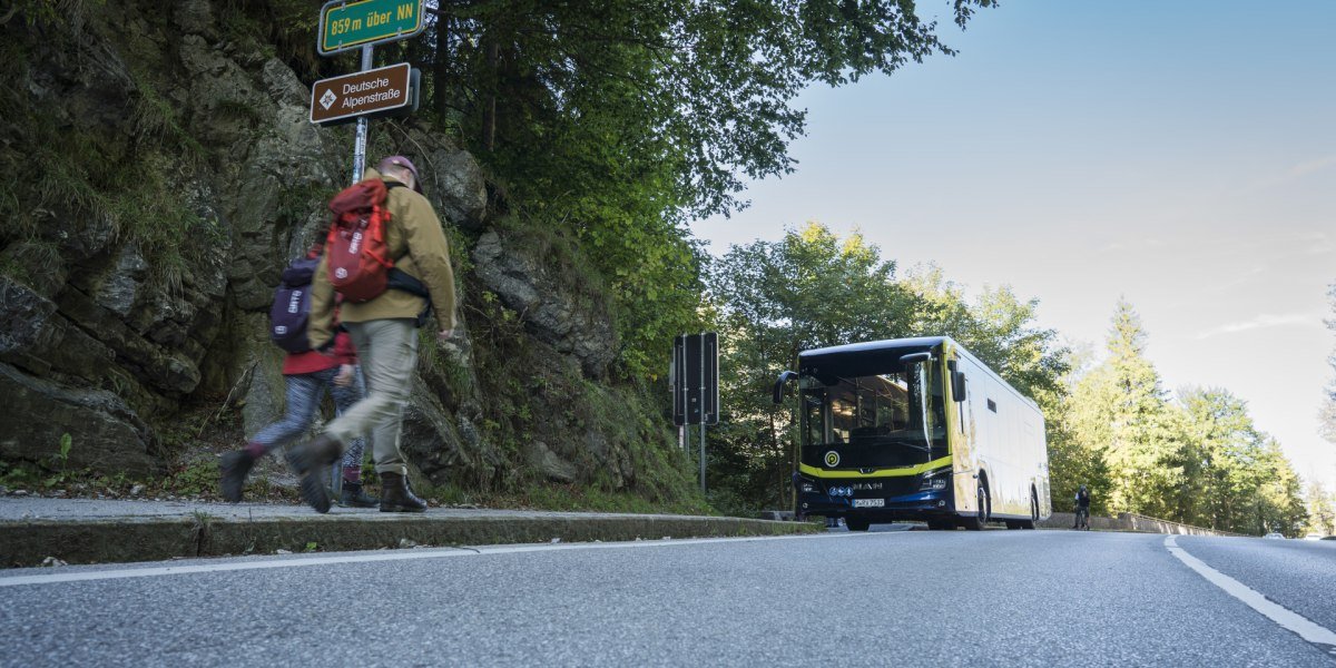 Zwei Wanderer mit Rucks&auml;cken gehen am Berg entlang, rechts h&auml;lt ein moderner Bus an der Stra&szlig;e des Kesselbergs, dar&uuml;ber Schilder mit H&ouml;henangabe und Hinweis auf die Deutsche Alpenstra&szlig;e., &copy; Landratsamt Bad T&ouml;lz-Wolfratshausen|Dietmar Denger